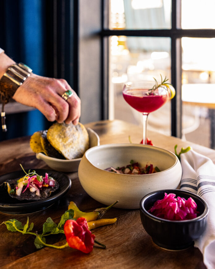 A person picks up a corn tortilla amongst a table of various small dishes holding ceviche and ingredients, plus a cocktail sitting to the back right.