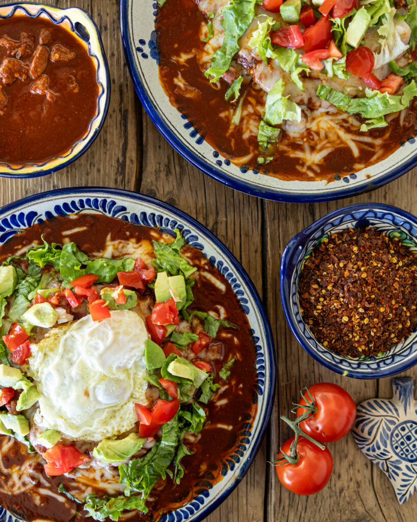 Freshly made Red Chile and Blue Corn Enchiladas by Carmella Padilla sit on a table with a side of beans, salsa, and tomatoes nearby.