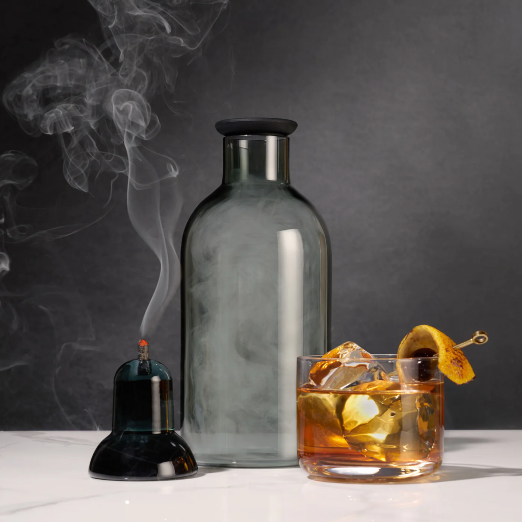 A smoked grey bottle sits in front of an old fashioned cocktail with smoke rising up through the photo.