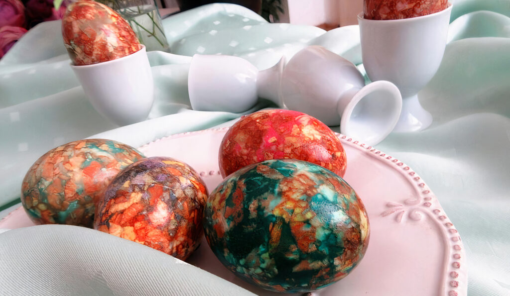 Four marble colorful dyed Easter eggs sit on a pink plate surrounded by silk and egg cups.