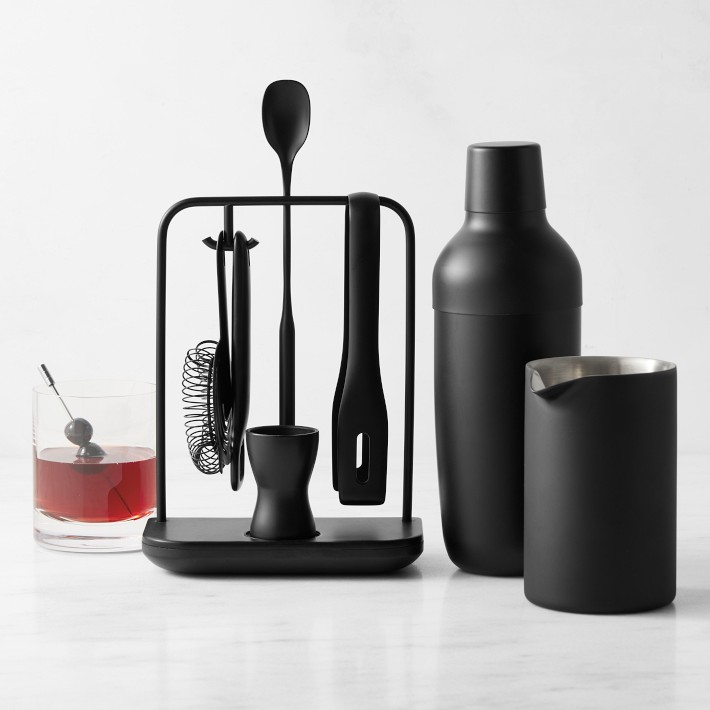 A black barware set sits beside a cocktail featuring a cocktail shaker, stirring spoon, strainer, and more.