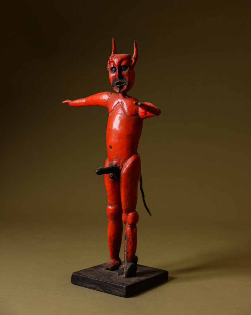 A statue of a red devil completely naked with a greenish brown background in the vaults at the Museum of International Folk Art.