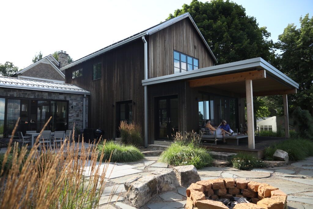 The outside of a wooden Chautauqua lake house with a fire pit outside and two people sitting on the porch,.