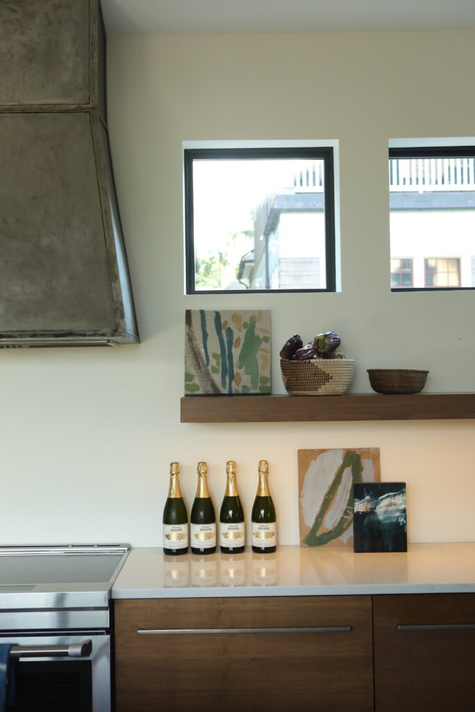 The kitchen counter of a Chautauqua lake house holds bottles of wine, paintings, and bowls beside a stove and stove hood.