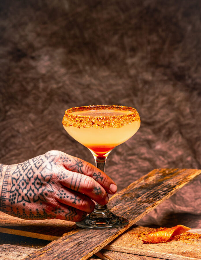 A cocktail glass with an orange spring cocktail is help in the tattooed hand of a New Mexico mixologist.