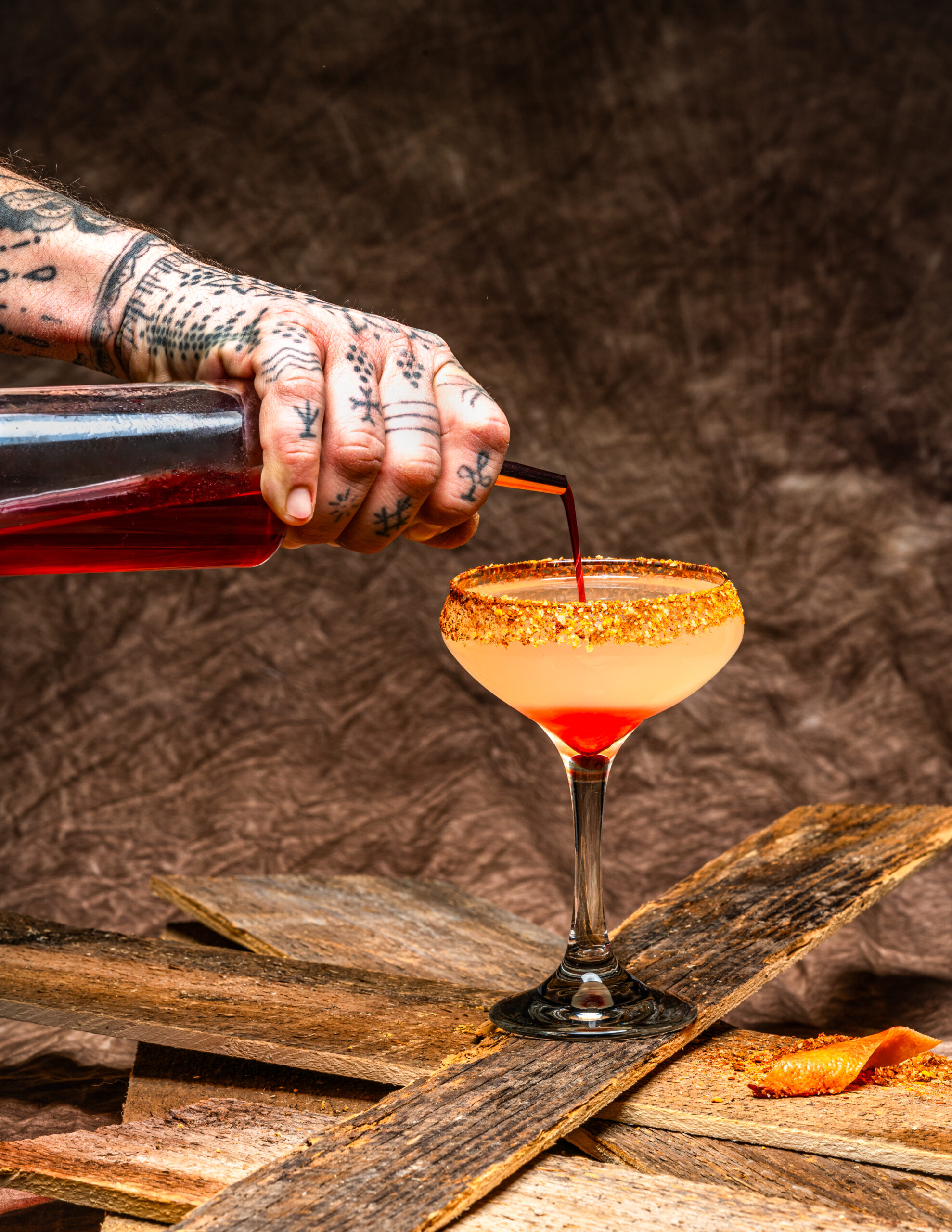 A cocktail glass holds an orange drink as a bartender pours a red liquid into the glass from above.
