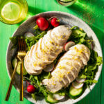 A white plate holds a salad arranged with tomatoes and sliced chamomile ginger poached chicken on top with two gold forks off to the side. It all sits against a green background.