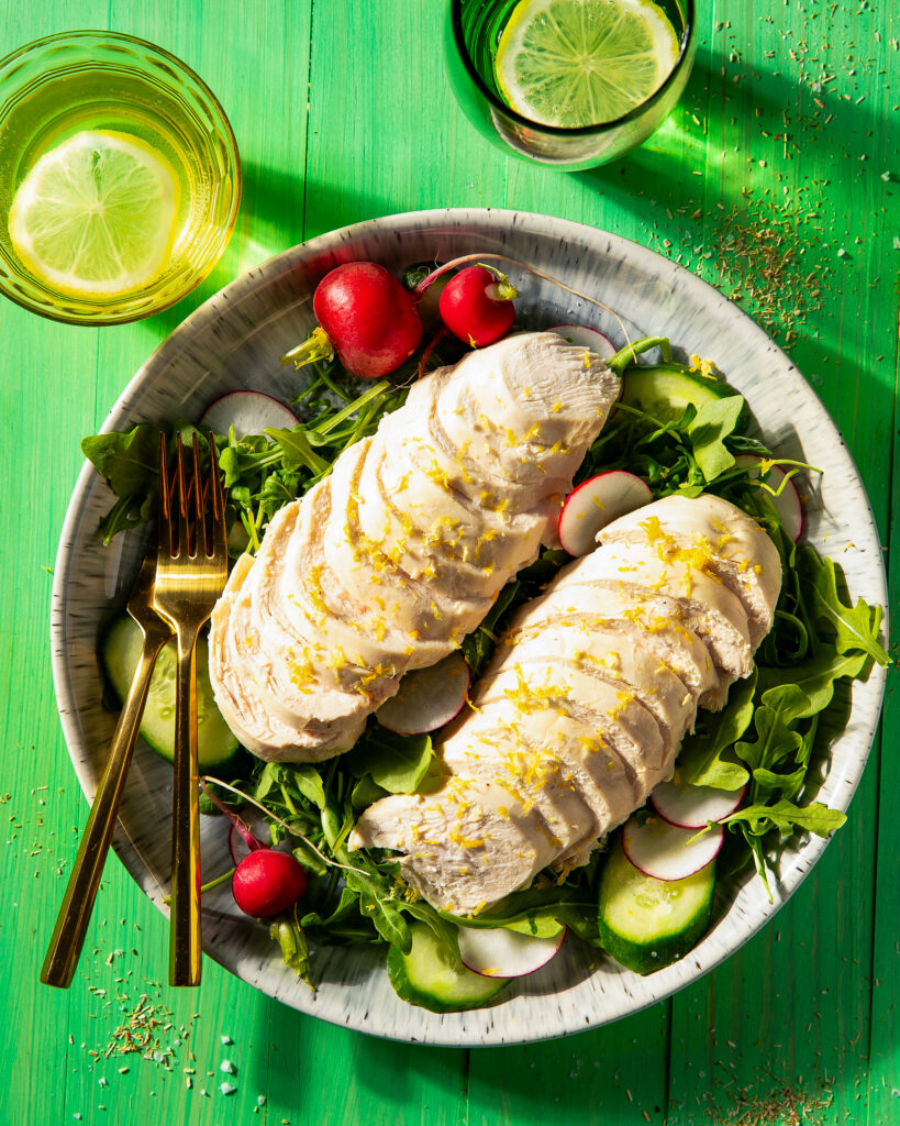 A white plate holds a salad arranged with tomatoes and sliced chamomile ginger poached chicken on top with two gold forks off to the side. It all sits against a green background.