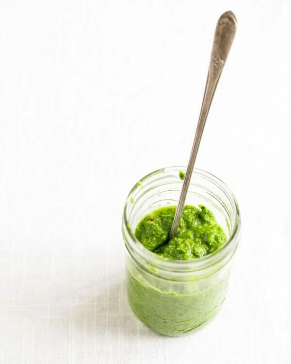 A glass jar holds a green ramp chimichurri sauce with a spoon sitcking out of it, all against a white background.