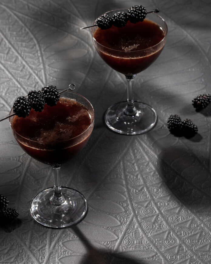Two Back to Black cocktails in a deep brown color with a pick of blackberries on top of each glass, all on a textured grey surface.
