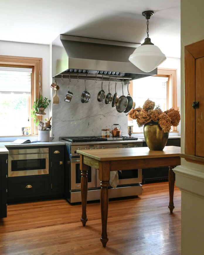 The silver stove, oven, hood, and sink sit against the wall of a remolded kitchen, a wooden table sits in the middle of the room with orange flowers on it.