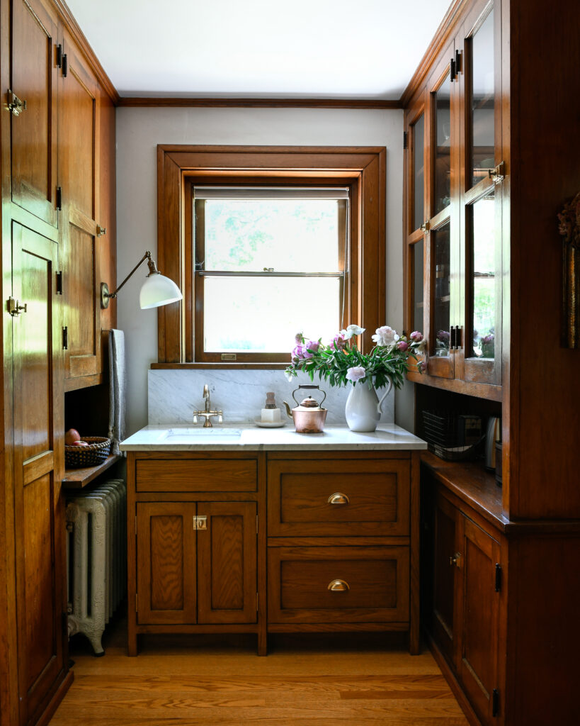 A sink with wooden cabinets sits between two taler wooden cabinets with a small window above the white sink and lamp.