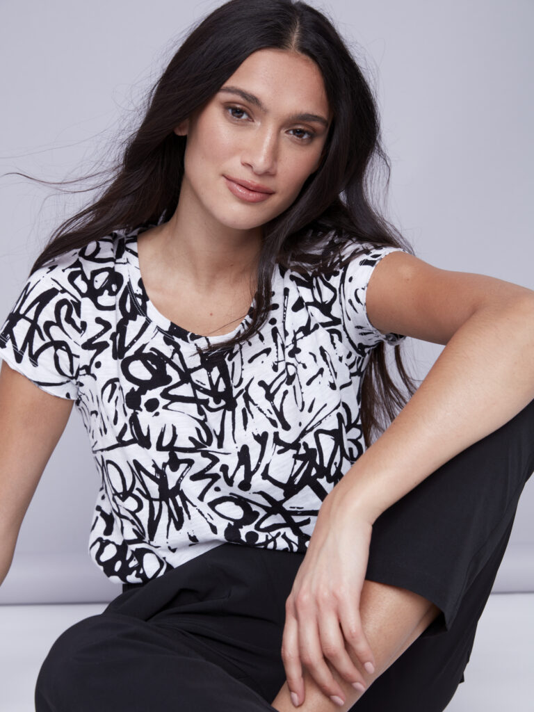 A woman sits on the floor in a black and white pattern shirt and black pants, her arm across her knee.