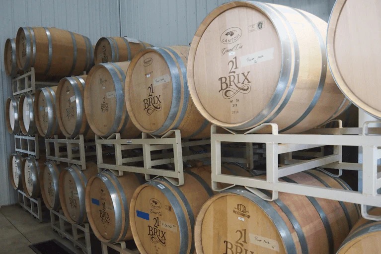 Light tan colored wine barrels from 21 Brix stacked sideways on metal racks.