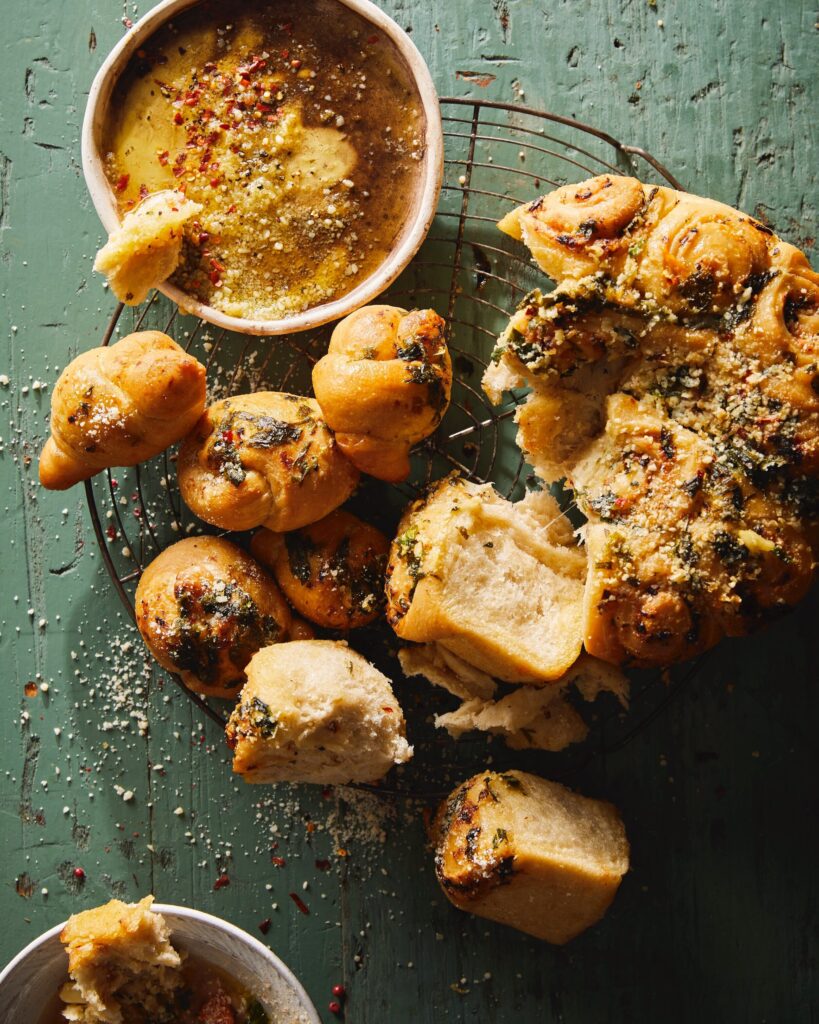A plate of garlic knots sit on a green table top beside a side dish of ramp butter.