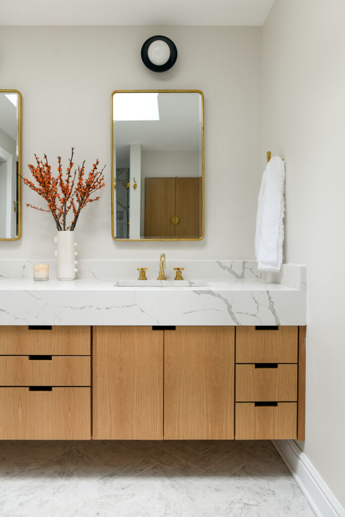 A bathroom marble sink with pale wood cabinets and a mirror above the sink is decorated with gold embellishments on the faucet and mirror. 