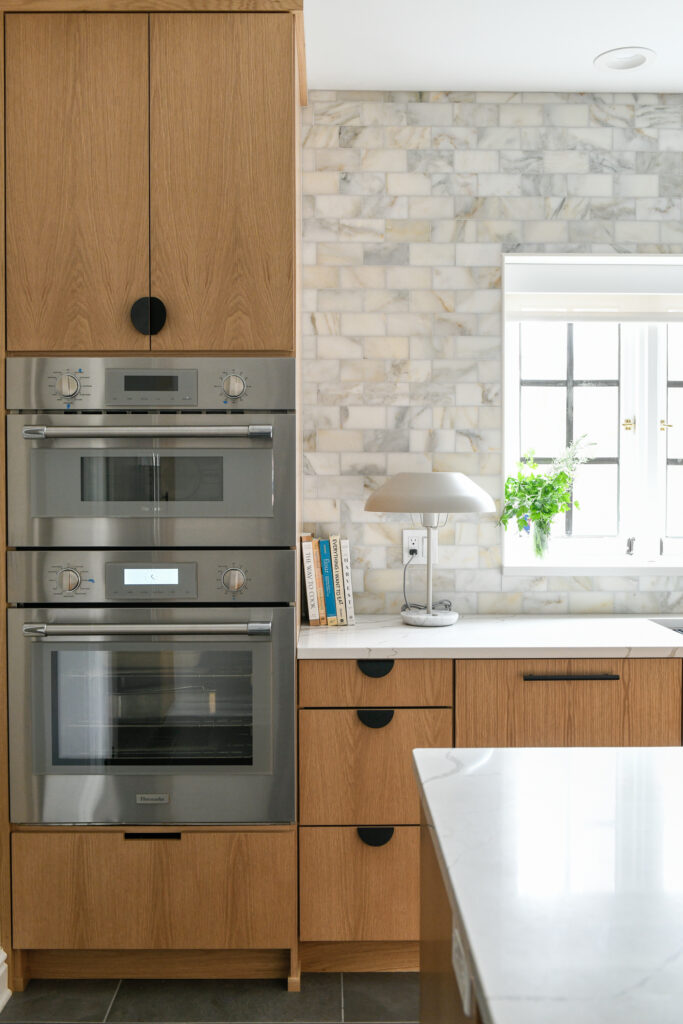 A black kitchen oven built into wood cabinetry in a kitchen as a lamp and books sit on the countertop beside it. 