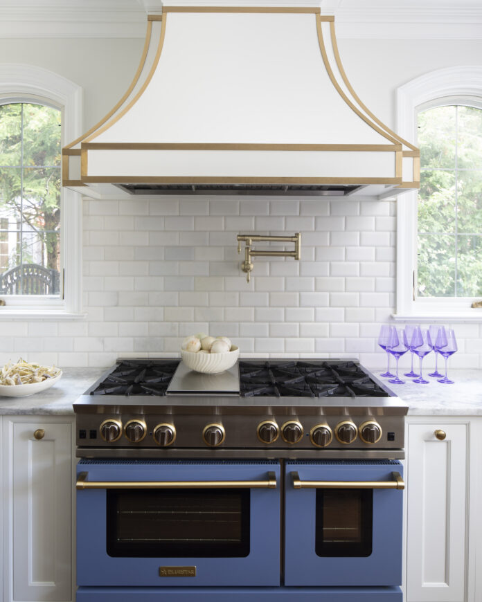 A matte blue stove with black embellishments sits below a white stove hood with gold embellishments. Purple wine glasses sit to the right of the stove on a white countertop.