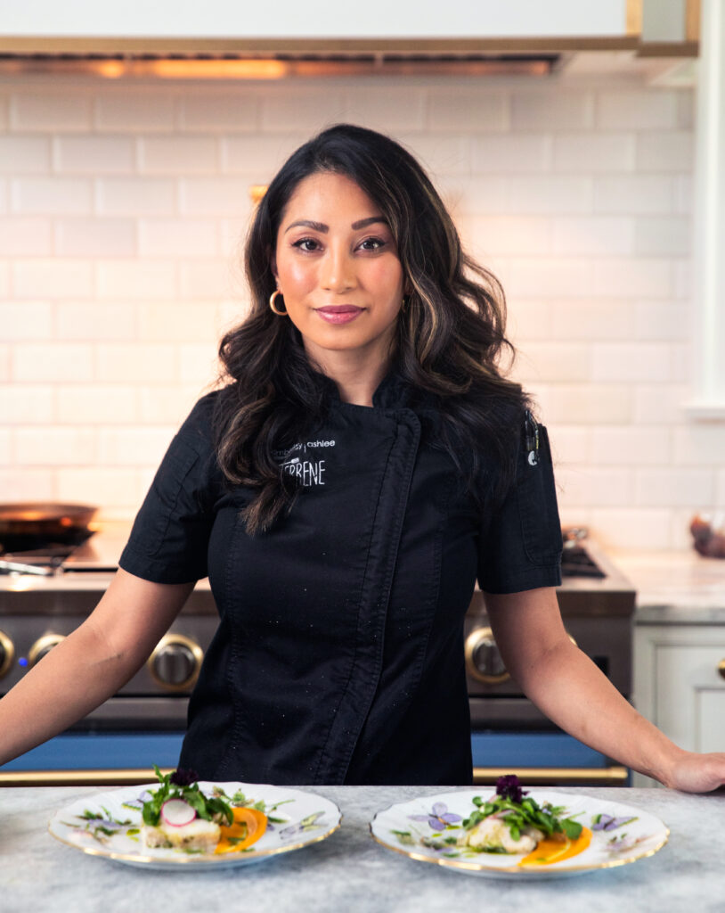 A woman in a black chefs shirt stands at a counter with two white plates in front of her.