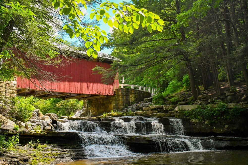 Packsaddle Covered Bridge, a red wooden bridge over small waterfalls and a clear stream in a wooded area.