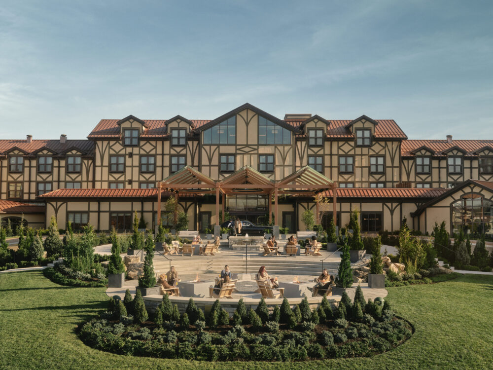 An outside image of The Grand Lodge at Nemacolin, a tan and brown building with terra cotta roofing and a large patio area with lush greenery in the front of the building.