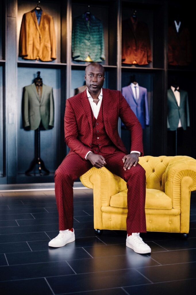 A man in a red suit sits on a yellow chair with other suits in the wall blocks behind him.