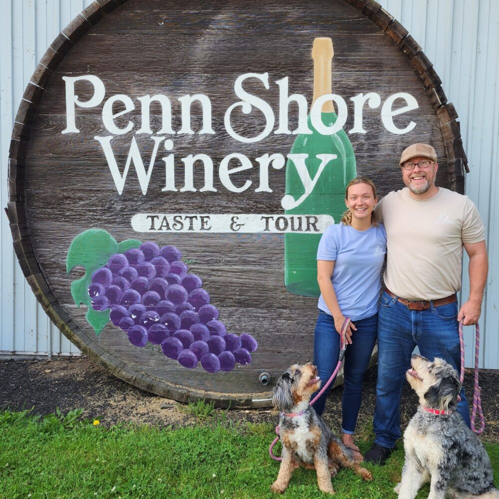 A man and a women standing in front of the Penn Shore Winery Sign with 2 dogs on leashes.