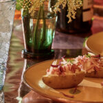 A yellow plate holds small potatoes with Pickled Ramps on top, a spritz drink to the left, and flowers behind the plate.