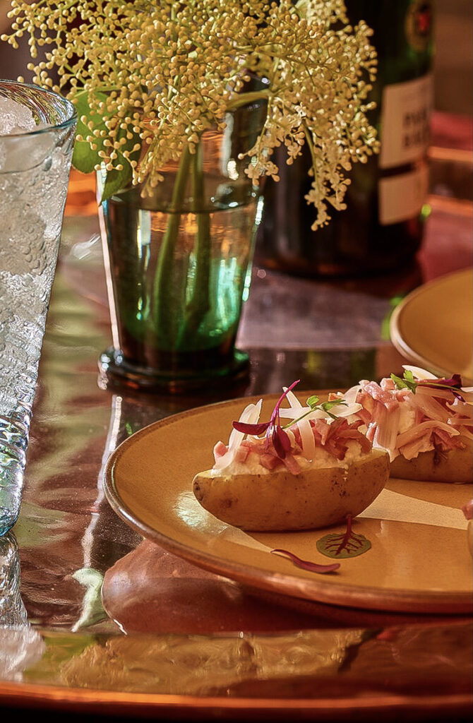 A yellow plate holds small potatoes with Pickled Ramps on top, a spritz drink to the left, and flowers behind the plate.