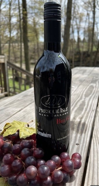 A bottle of red wine with grapes in front of it sitting on a wooden picnic table outside in what looks like a wooded area.