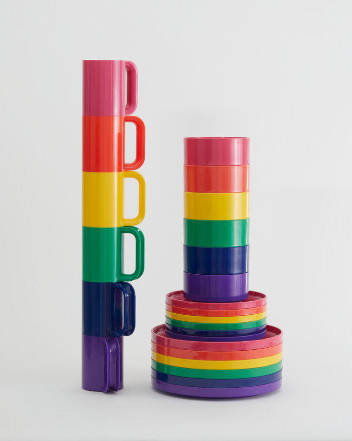 A set of rainbow mugs are stacked on top of each other to the left of a stack of rainbow Hellerware dinner plates and bowls.