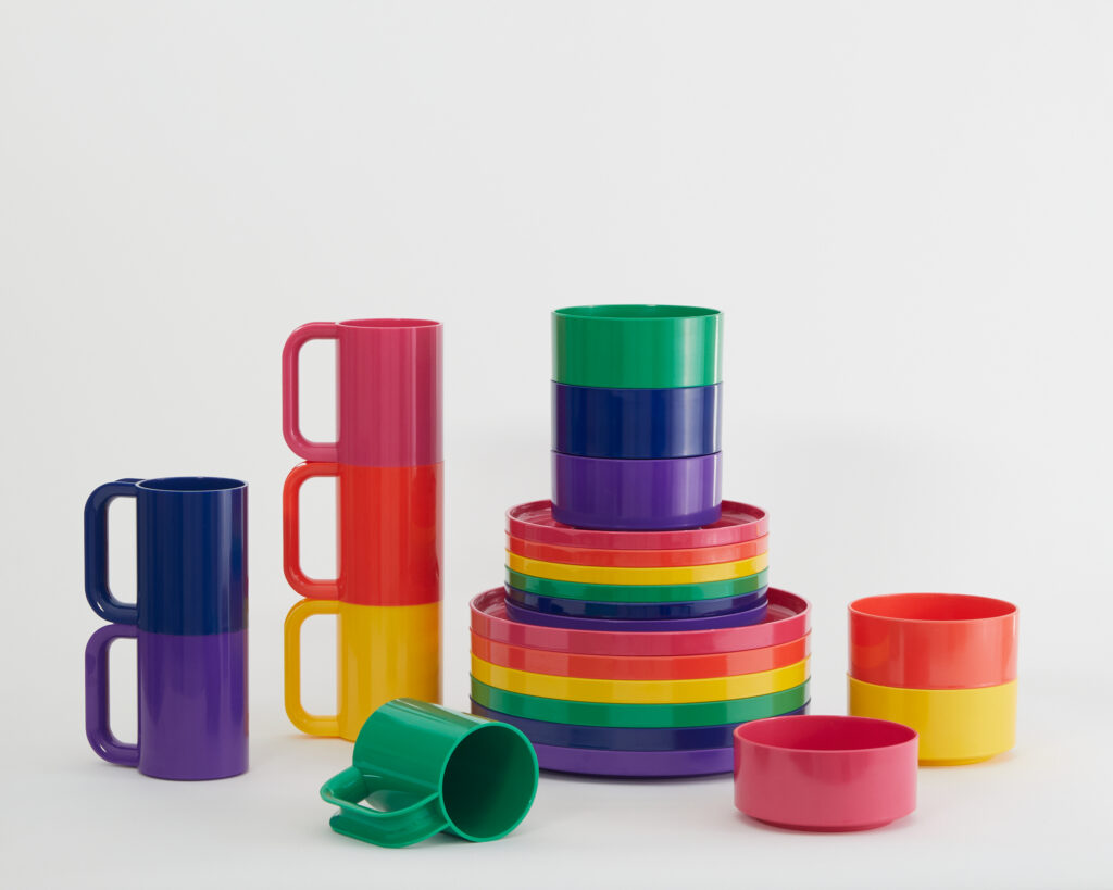 Two stacks of rainbow mugs from Hellerware sit beside a stack of rainbow plates and bowls while a green mug and red bowl sit in front of the piles.