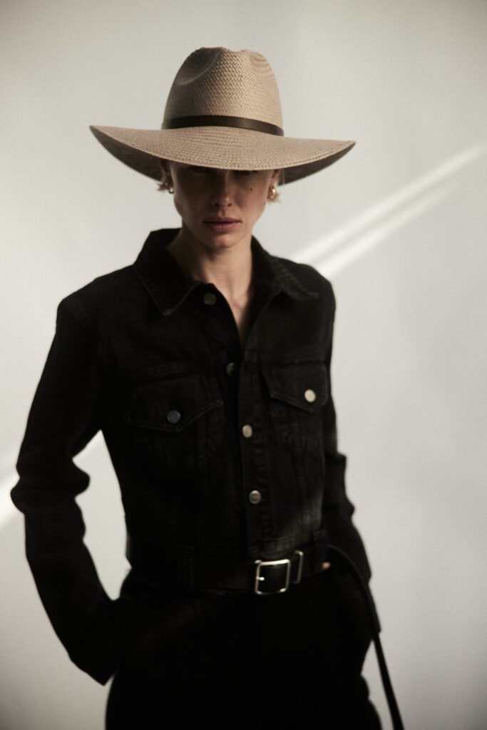 A woman in an all black jacket and beige cowboy hat stands in front of a neutral background, one hand in her pocket.