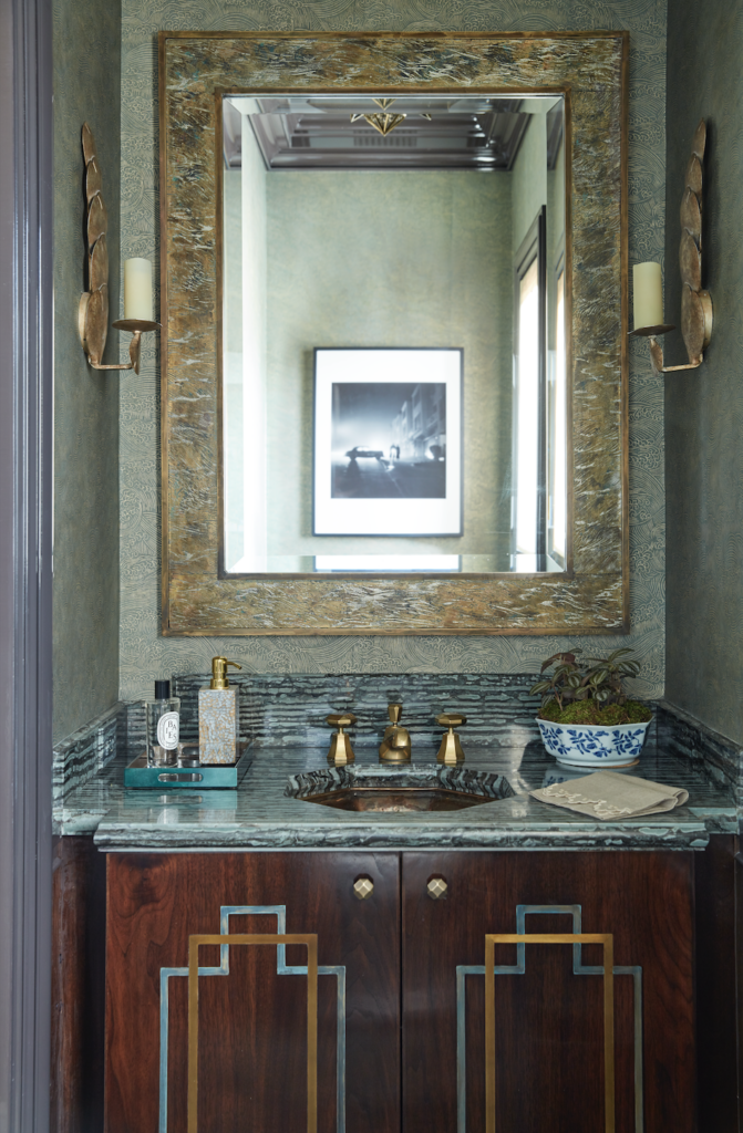 A dull sage green paint on a wall in a bathroom reflecting in the bold mirror above the marble sink.