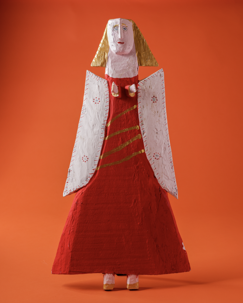 A handcrafted angel in a red dress with white wings at the museum of international folk art sits against a bright orange background.