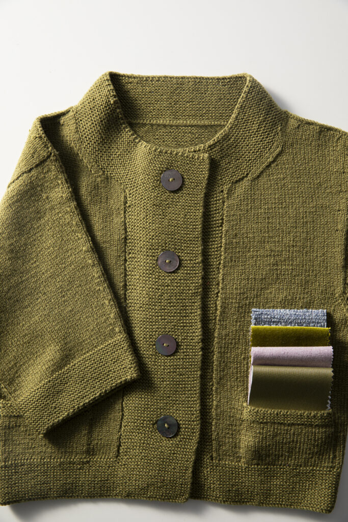 A green sweater with big buttons and fabric swatches sits on a white background.