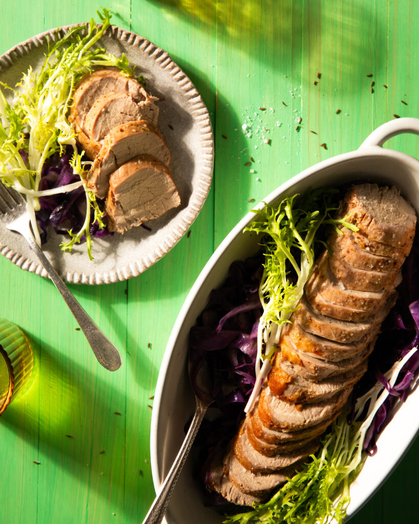 A white dish and beige plate both hold slices of toasted caraway pork tenderloin accompanied by greens and silverware all on a green background.