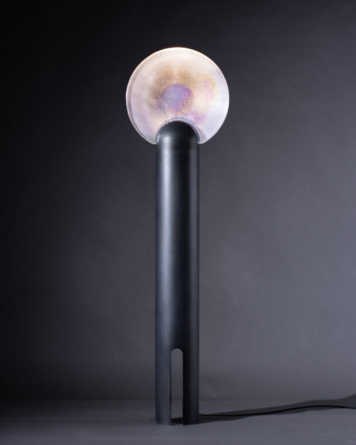 A round iridescent lamp sits on top of a long, wide black lamp post.