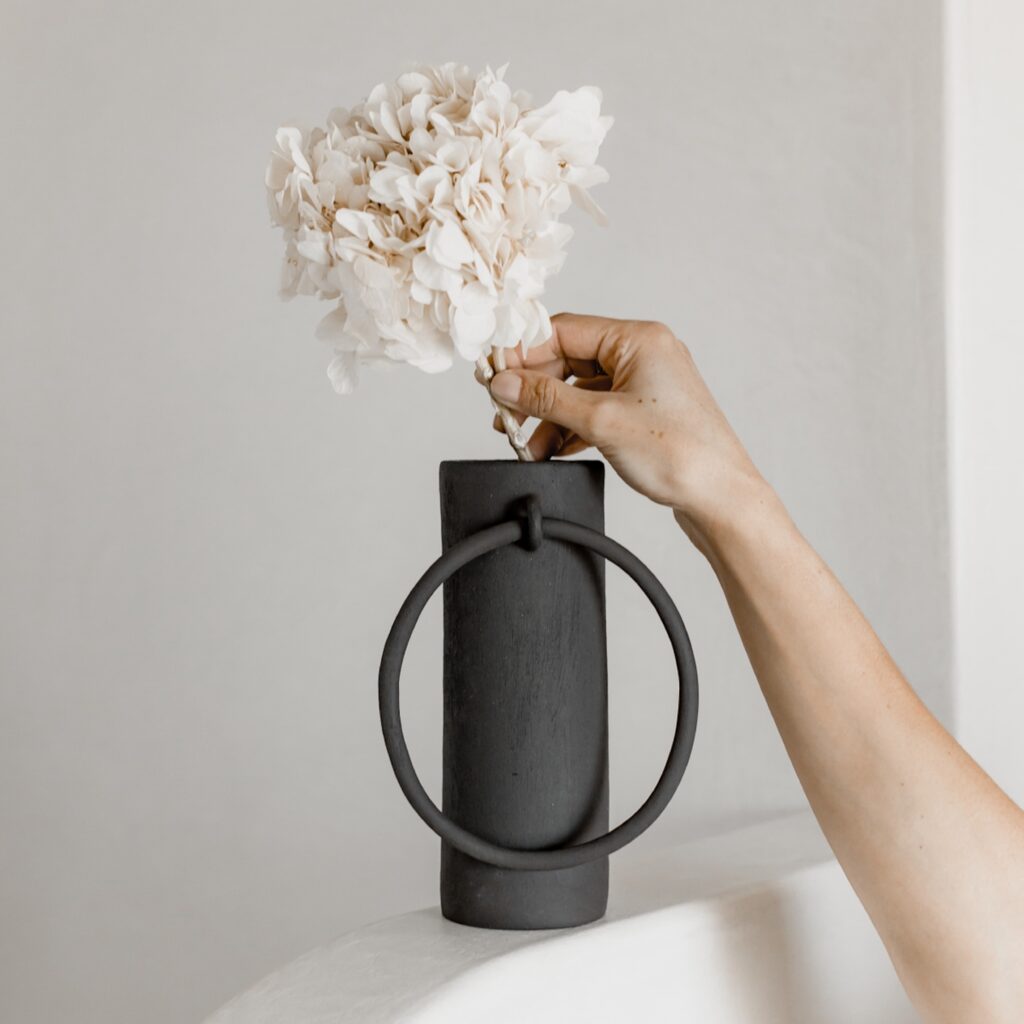A black vase with a circle ring holds white flowers as a woman's hand touches the flowers.
