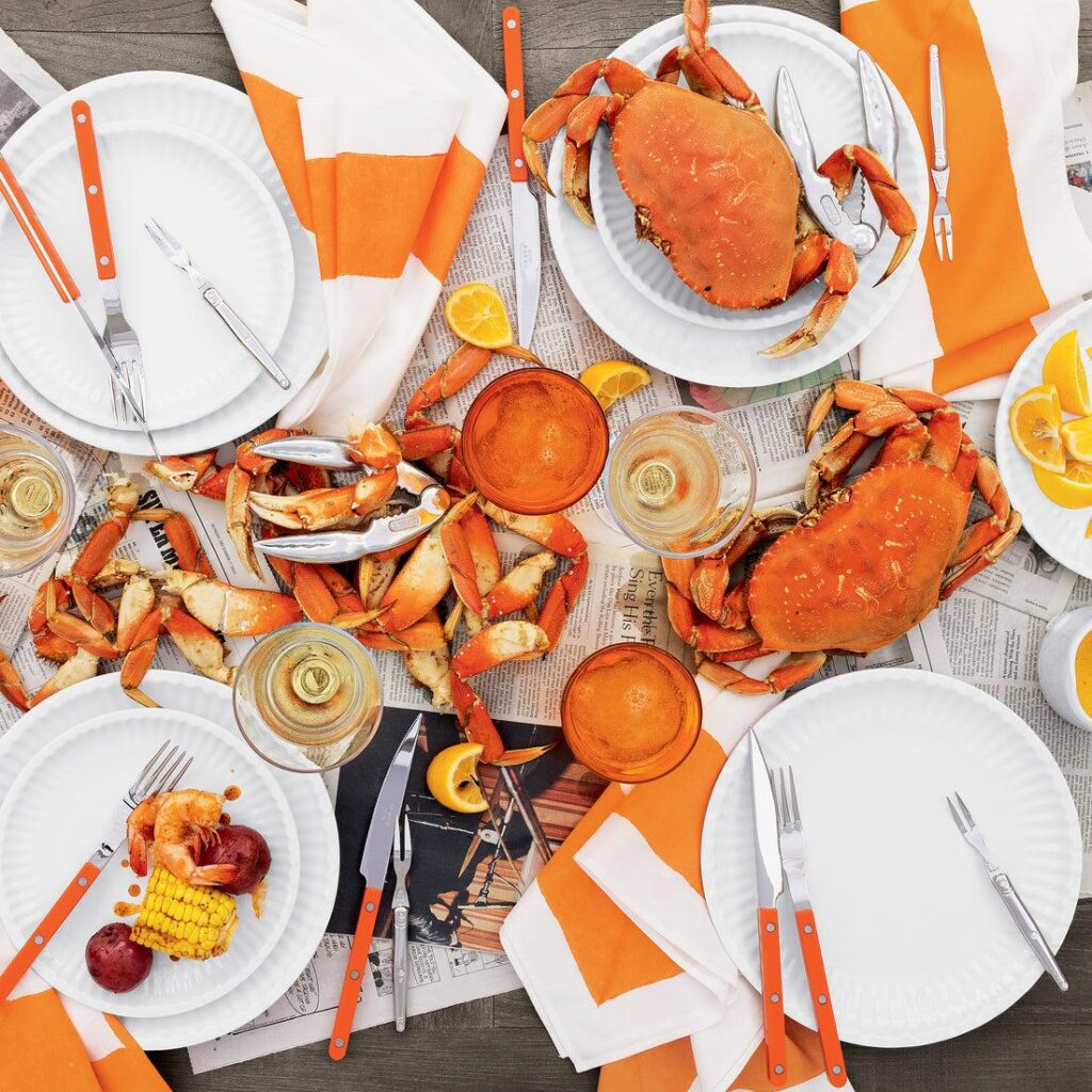 A variety of white tableware, white and orange napkins, orange accented silverware, and many crabs decorate a wooden table covered in newspaper while dining outdoors.