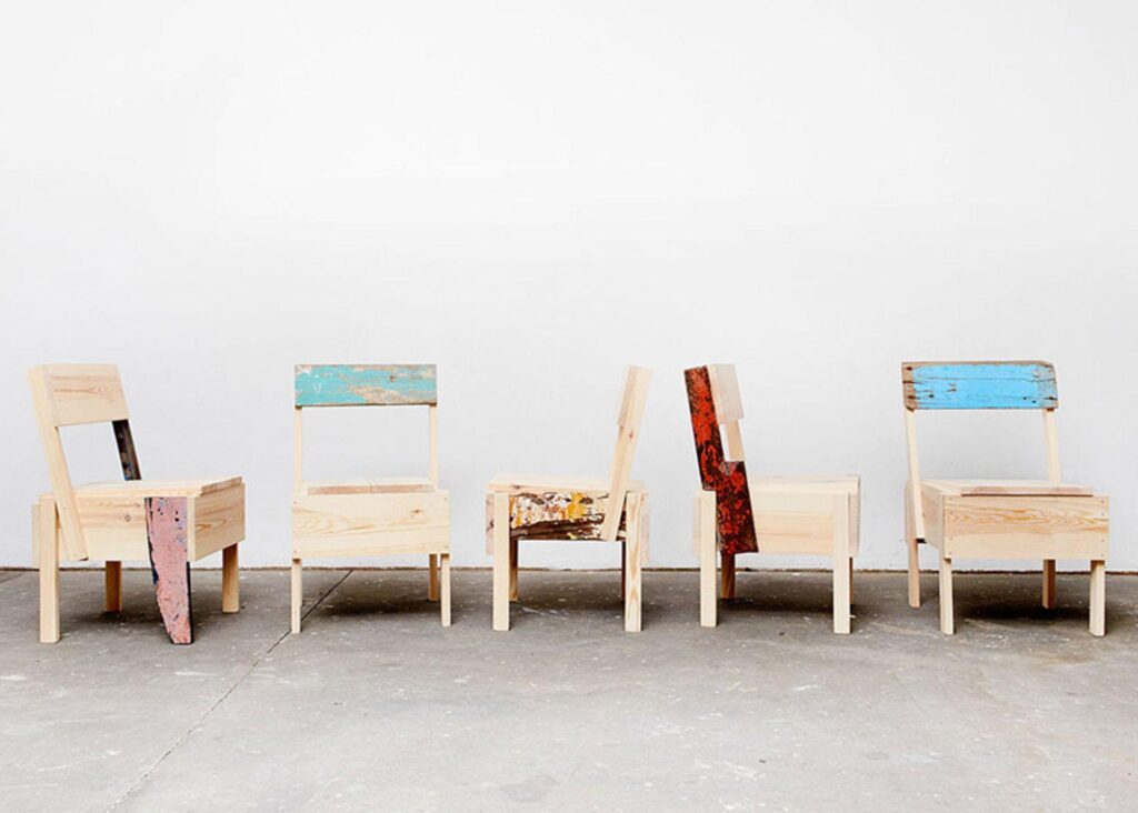 A series of 5 chairs made from bleached wood while each features a piece of colored metal as one of the limbs.
