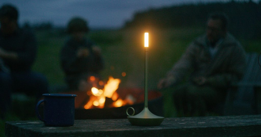 A small rechargeable camping lamp sits on a wooden bench beside a coffee cup while three campers sit around a fire in the background.