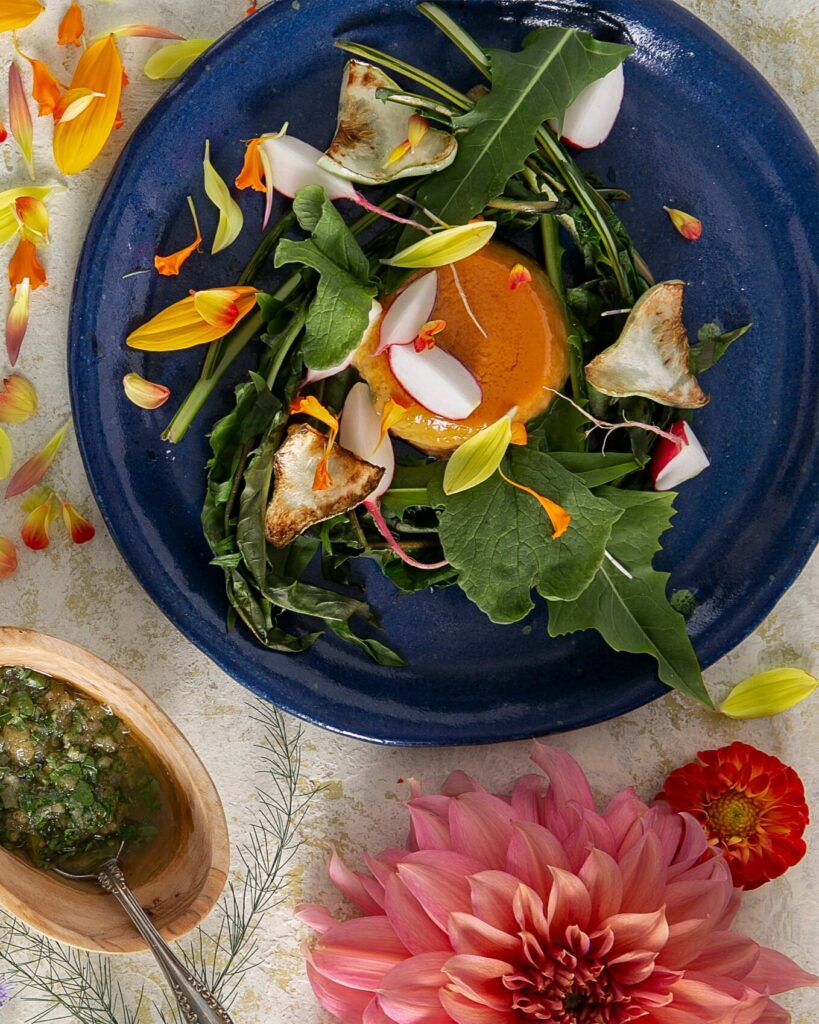 A blue plate holds Kohlrabi Flan with a salad of greens, mushrooms, ramps, and radishes all on a marble table with dandelions and other flowers around the plate.