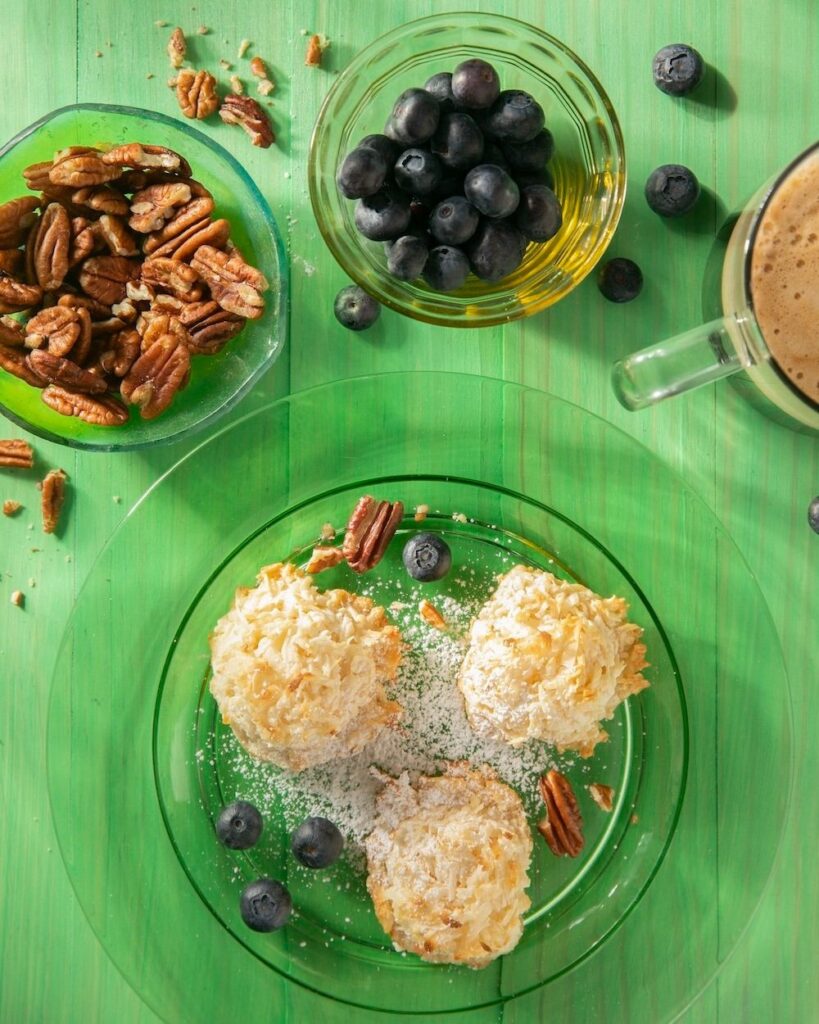 On a green table sits a plate with coconut macaroons, surrounded by walnuts and other topping bowls.