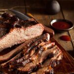A slab of smoked Passover Brisket sliced into pieces on a wooden table with a sauce sitting in the background.