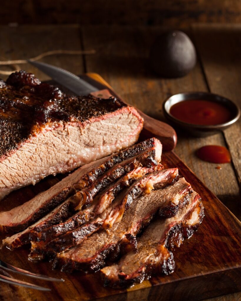 A slab of smoked Passover Brisket sliced into pieces on a wooden table with a sauce sitting in the background.