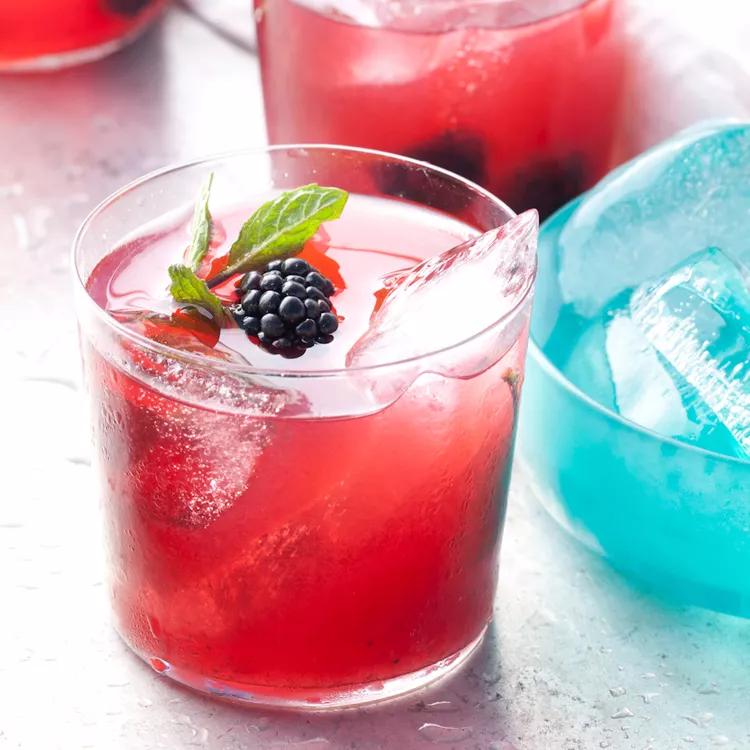A small glass holds red liquid with ice cubes, mint, and a blackberry floating throughout as a blue bowl of ice sits off to the right side.