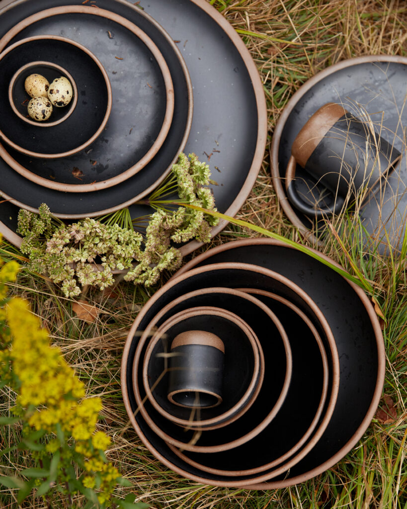 Three stacks of black repurposed plates sit on a table with spools of thread in the top bowl and foliage all around.