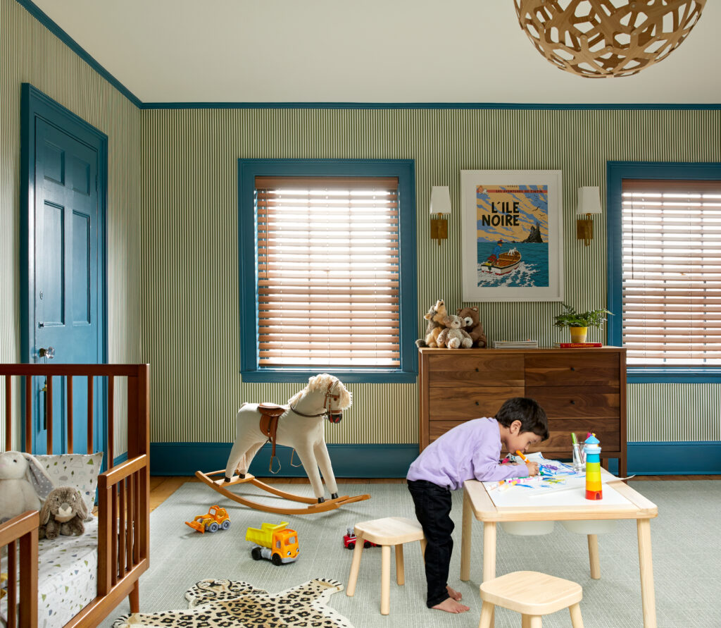 A child in a purple shirt draws on a table in his bedroom with patterned walls and blue trim around the windows and on the door.