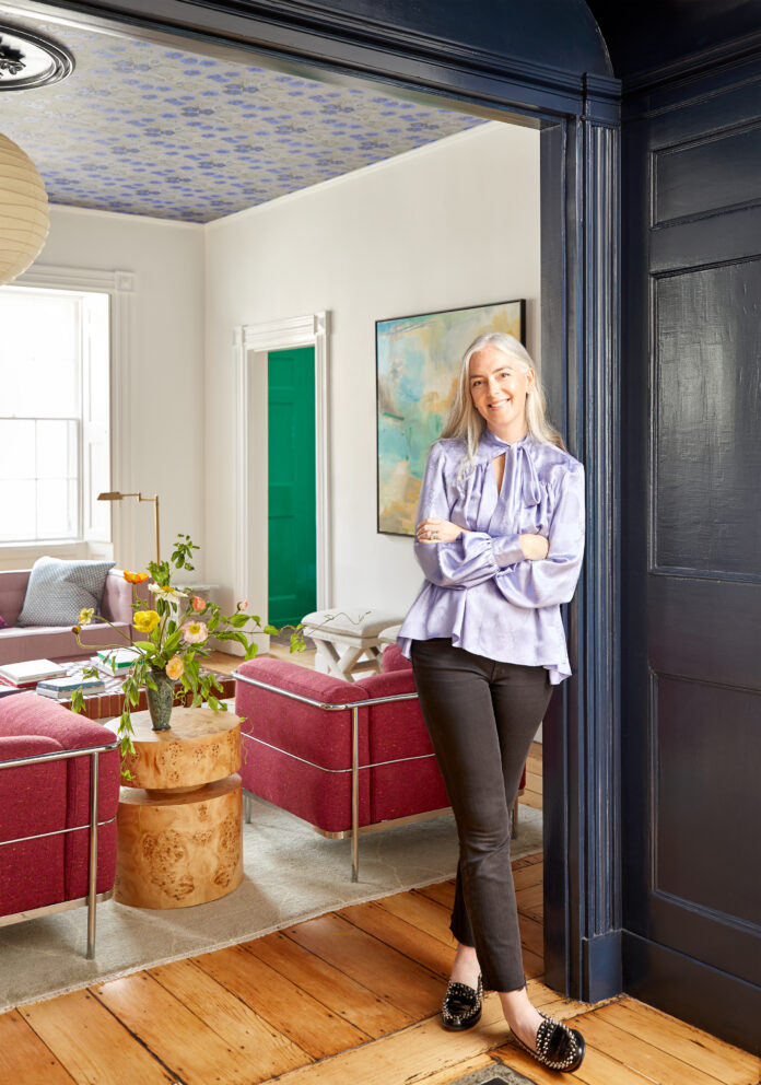 A photograph of Colleen Simonds in the home she designed with pink couches int eh living room behind her.